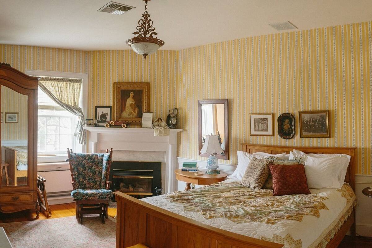 A bedroom with a wooden bed, a chair, a fireplace, and a mirror.