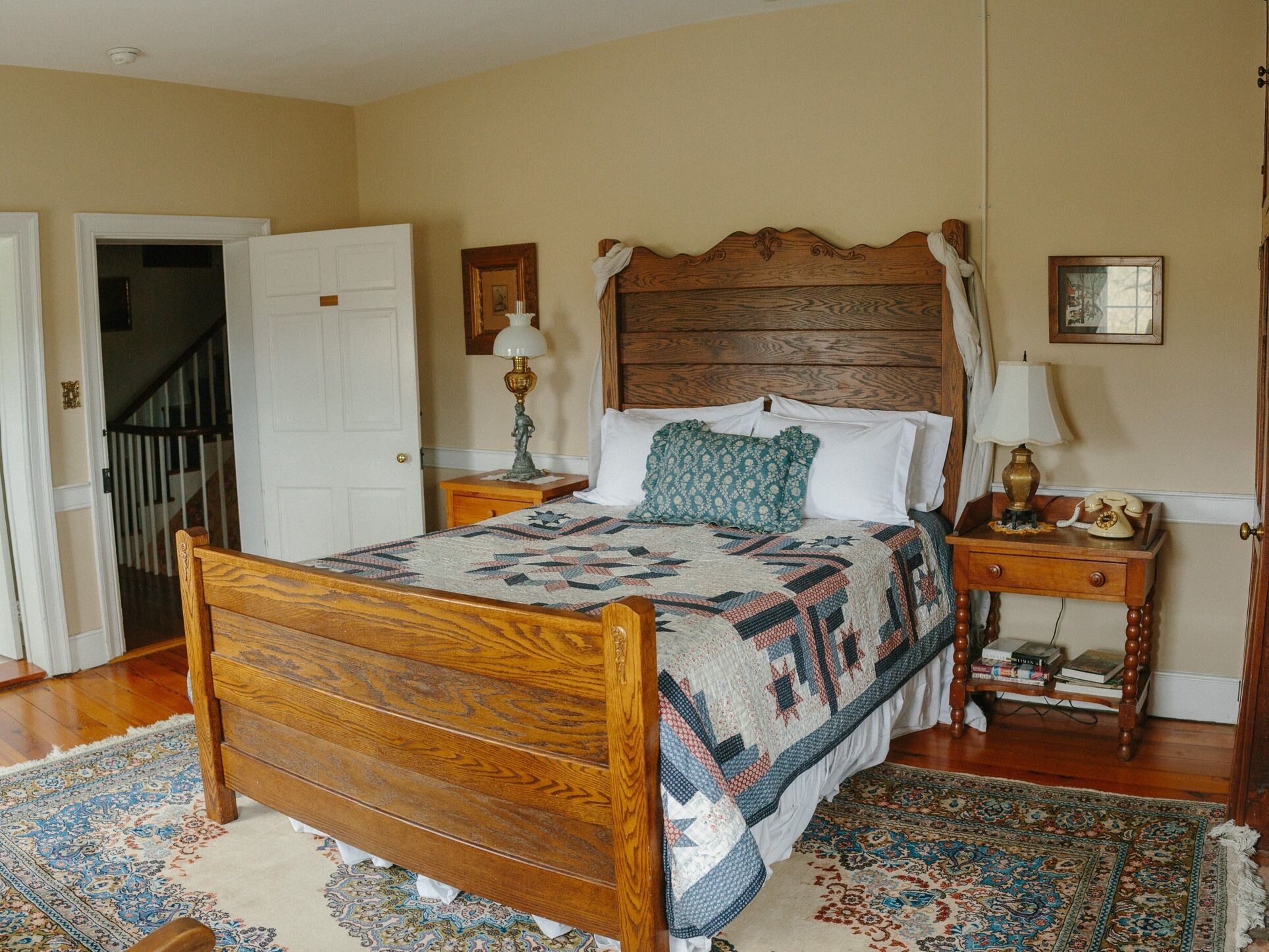 A bedroom with a wooden bed, a patterned quilt, a nightstand with a lamp, and a rug.