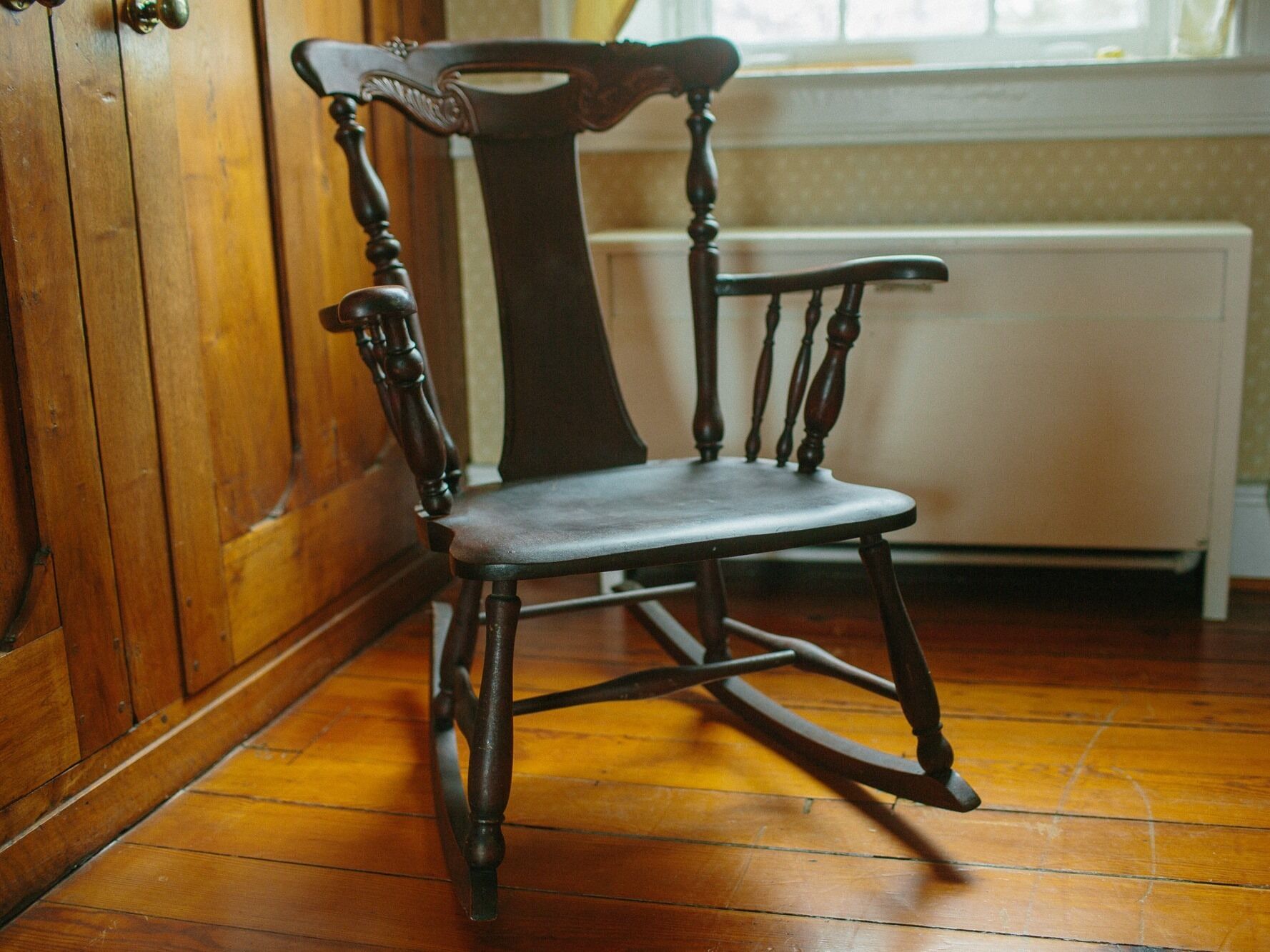 A wooden rocking chair with a high backrest and a cushioned seat, positioned in a room with wooden flooring and a radiator in the background.