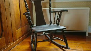 A wooden rocking chair with a high backrest and a cushioned seat, positioned in a room with wooden flooring and a radiator in the background.