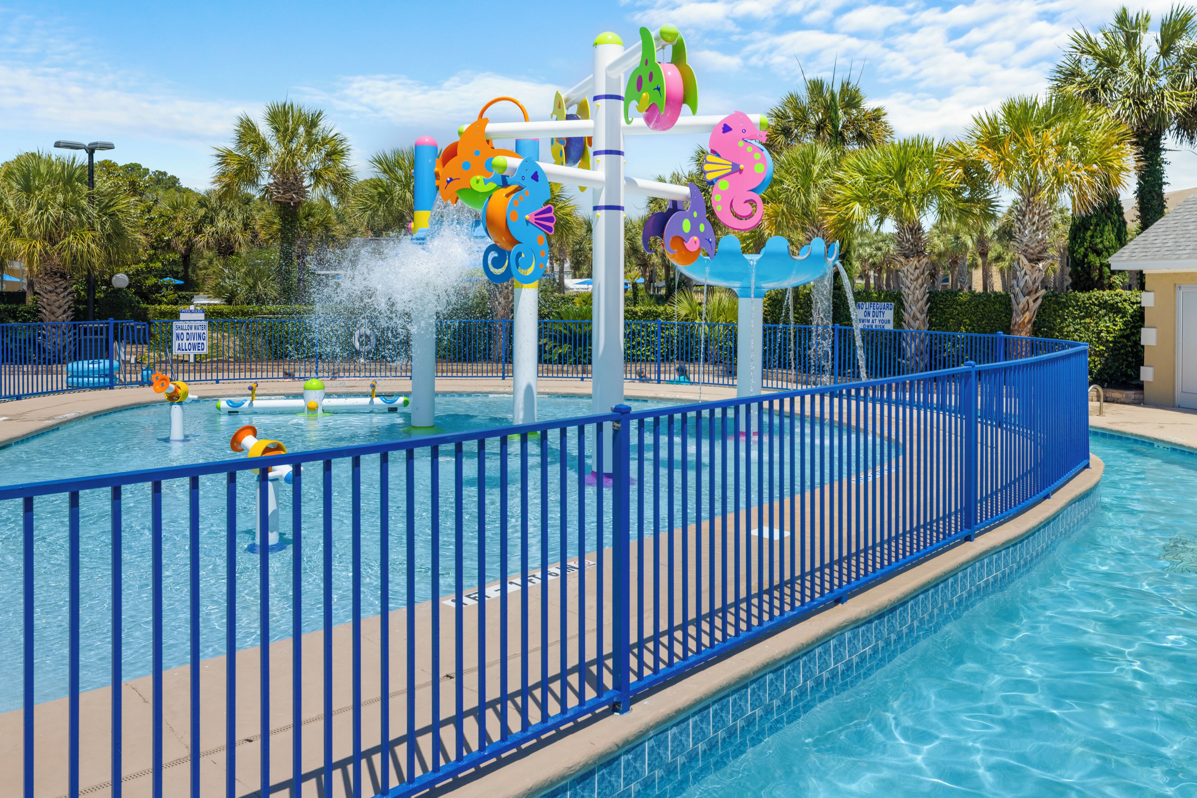 A swimming pool with a blue fence, water features, and palm trees in the background.