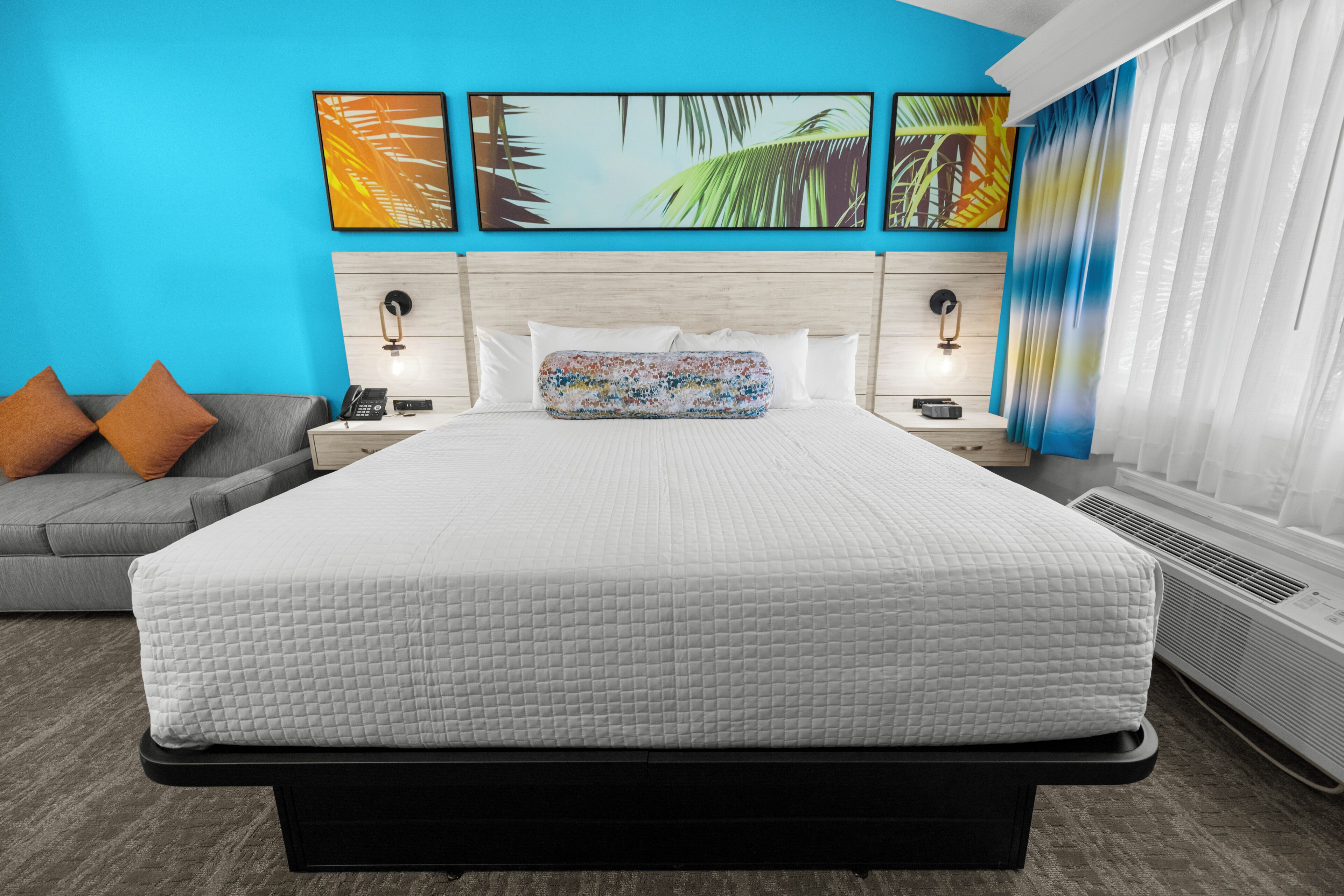 A hotel room with a large bed, a grey sofa, a nightstand with a phone, and wall art.