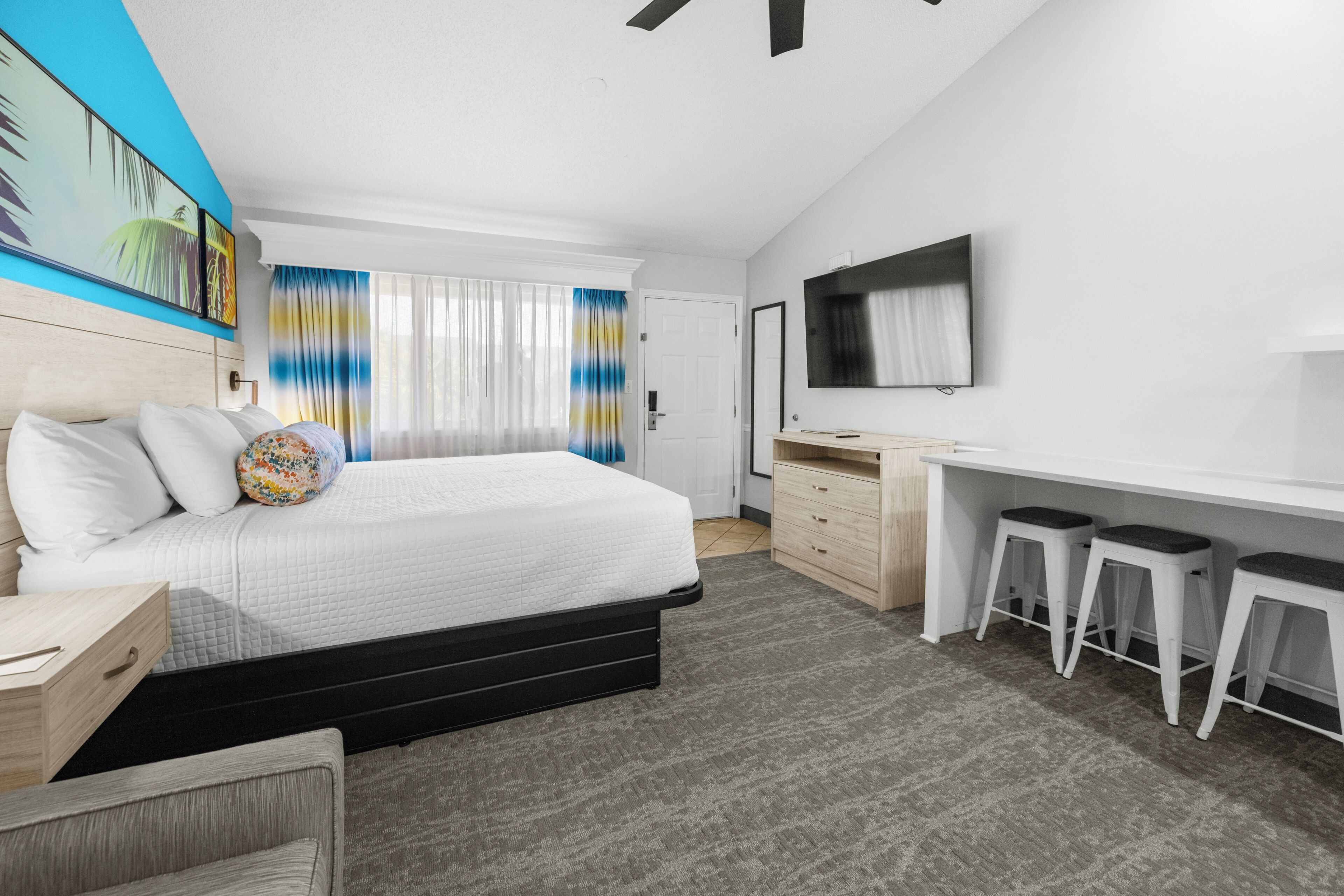 A modern hotel room with a bed, a desk, two bar stools, a TV, and a painting on the wall.