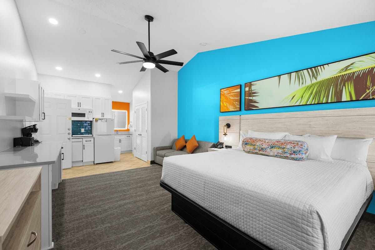 A modern hotel room with a bed, a ceiling fan, a kitchen area, and a bright blue accent wall.