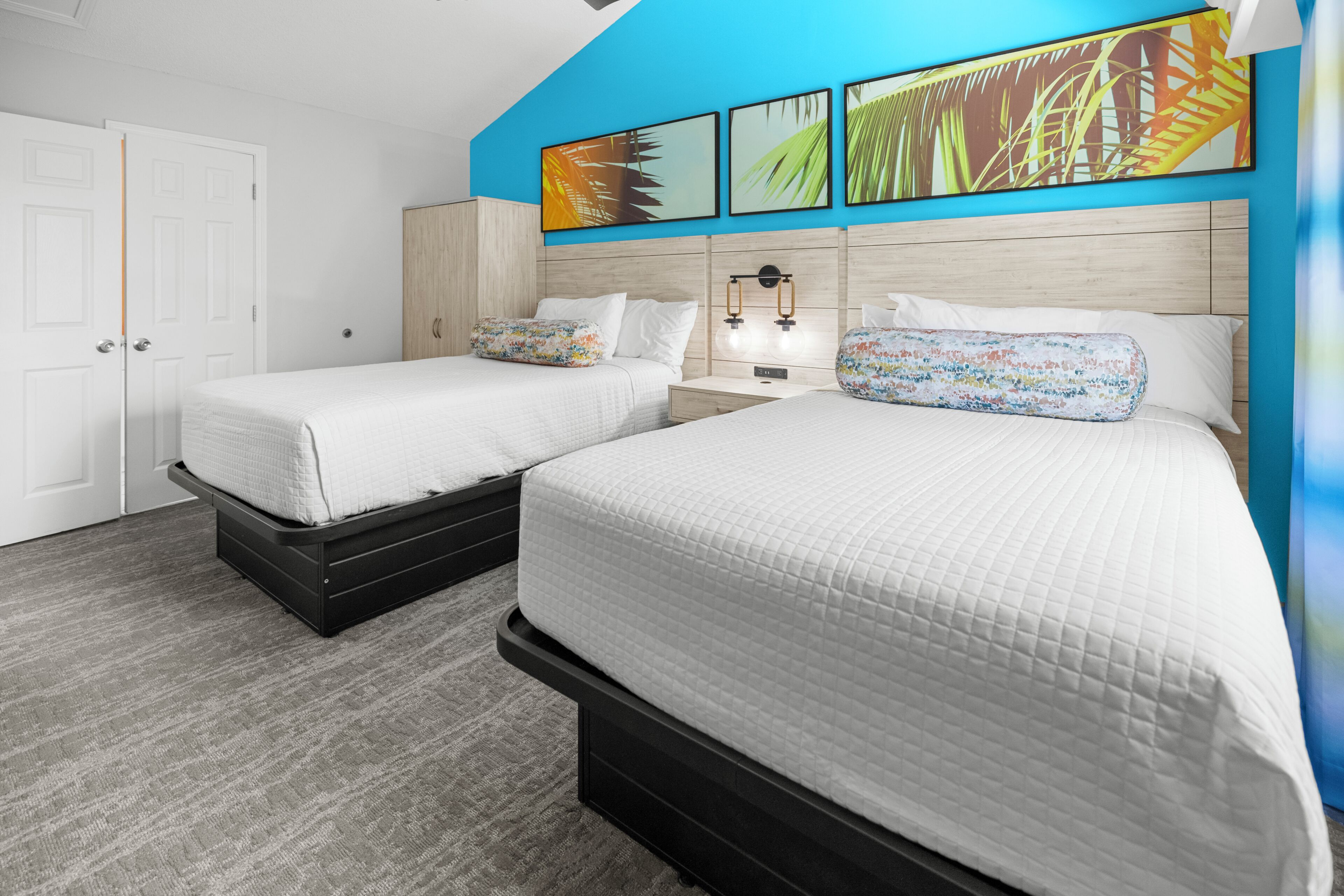 A hotel room with two beds, a wooden headboard, a nightstand, a wall-mounted lamp, and a door.