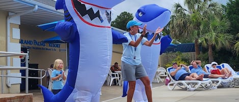 A person in a shark costume walking past a pool area with lounge chairs and a building labeled "Grand Palm's Resort".