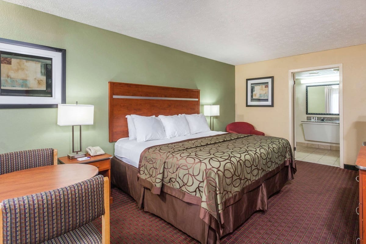 Une chambre d’hôtel avec un lit, un bureau avec une chaise, une petite table, une lampe, un miroir et une salle de bain visible à travers une porte ouverte.