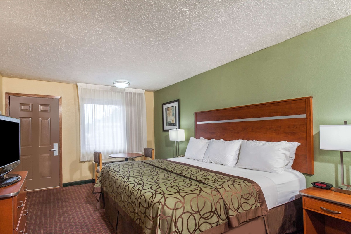 Une chambre d’hôtel avec un lit, un bureau avec une télévision, une chaise, une fenêtre avec des rideaux et un tableau encadré au mur.