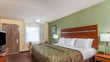 A hotel room with a bed, a desk with a television, a chair, a window with curtains, and a framed picture on the wall.