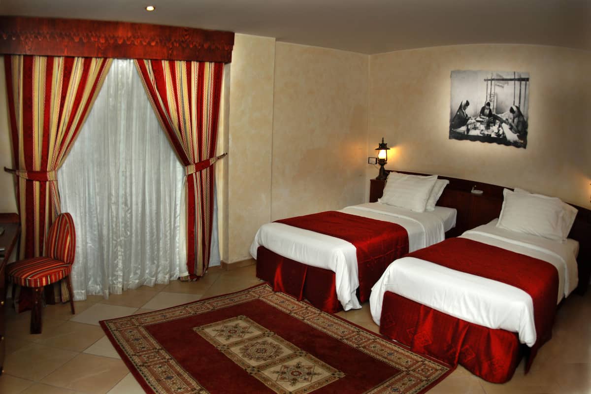 A hotel room with two beds, a chair, a large window with curtains, a wall-mounted lamp, and a framed picture.