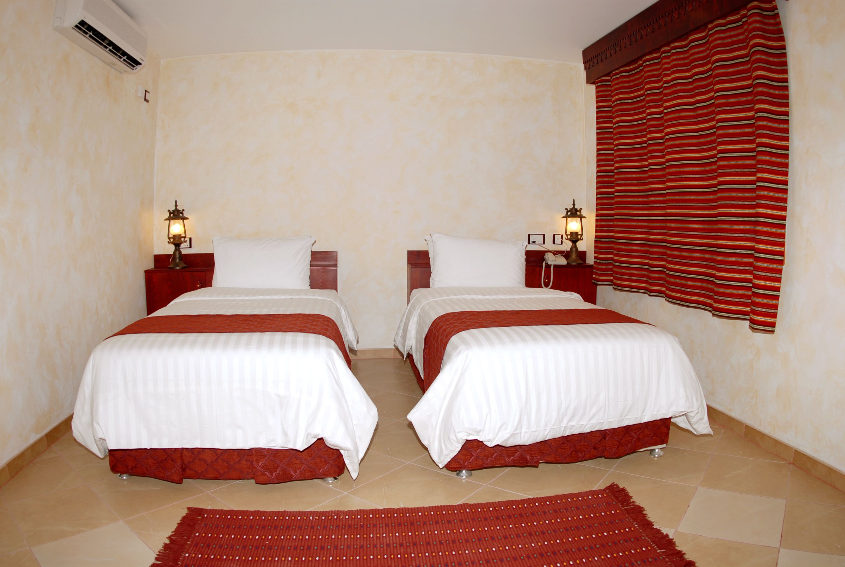 A hotel room with two beds, each with a red bedspread and white pillows, flanked by nightstands with lamps.