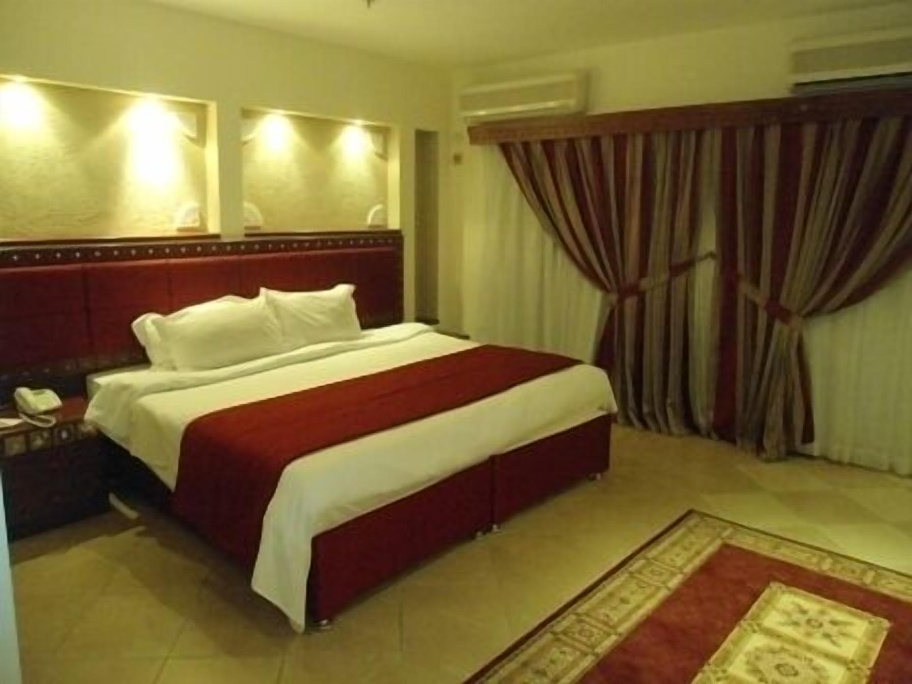 Habitación de hotel con una cama grande, una mesita de noche con lámpara, un teléfono y una colcha roja y blanca.
