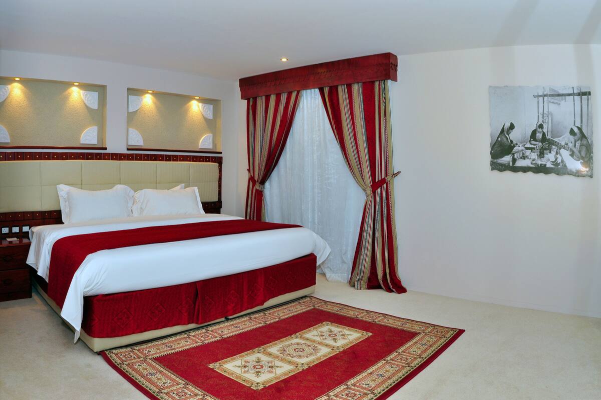 A hotel room with a large bed, a red and white bedspread, a red and white curtain, a red and white headboard, and a painting on the wall.
