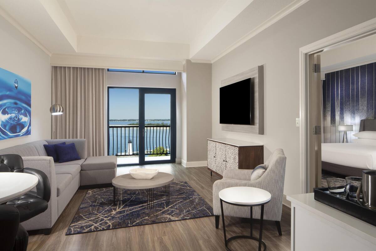 A modern hotel room with a balcony, a flat-screen TV, a dining area, and a sofa.