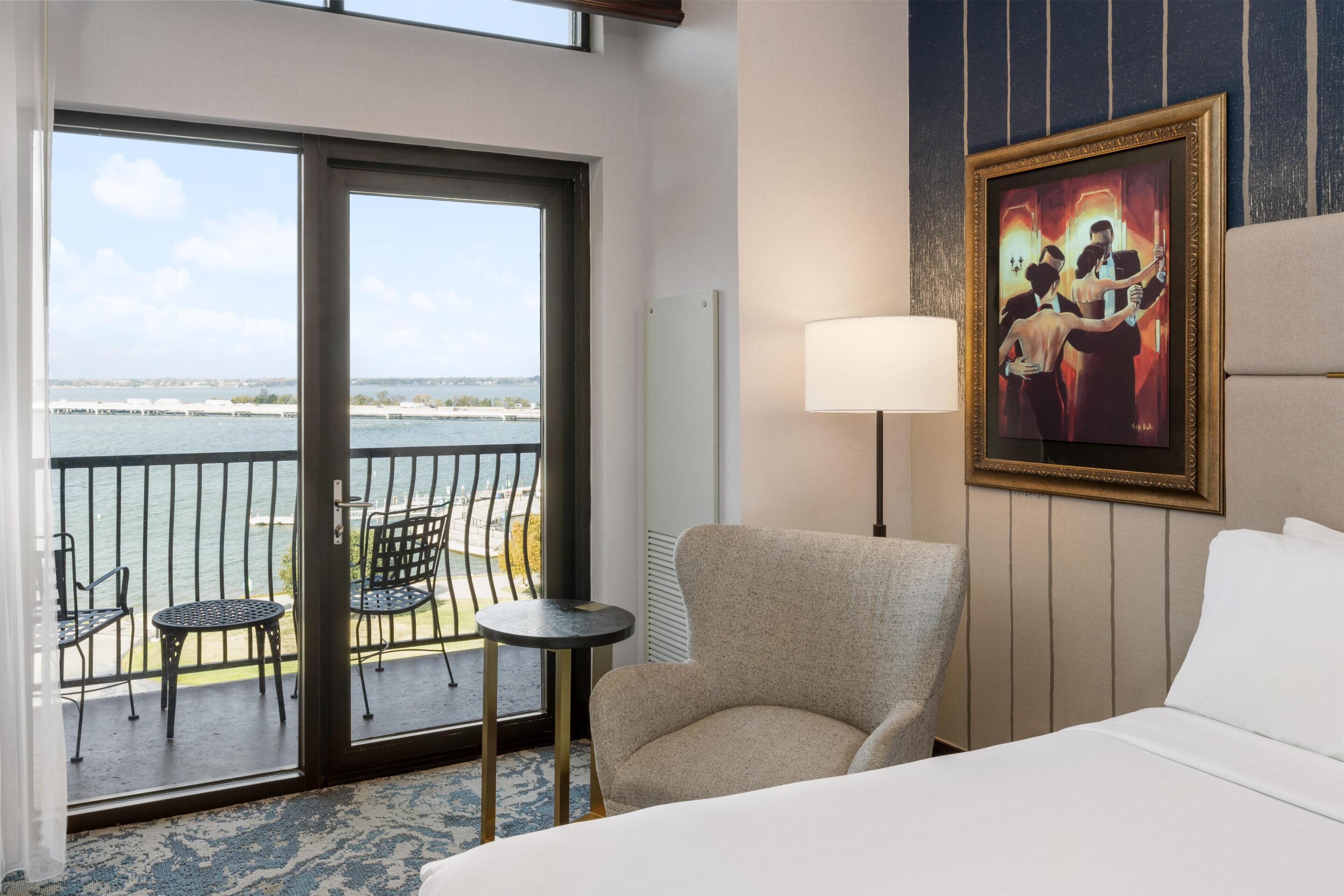 Habitación de hotel con cama, una silla, una mesita, un cuadro y un balcón con vista al agua.