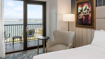 Habitación de hotel con cama, una silla, una mesita, un cuadro y un balcón con vista al agua.