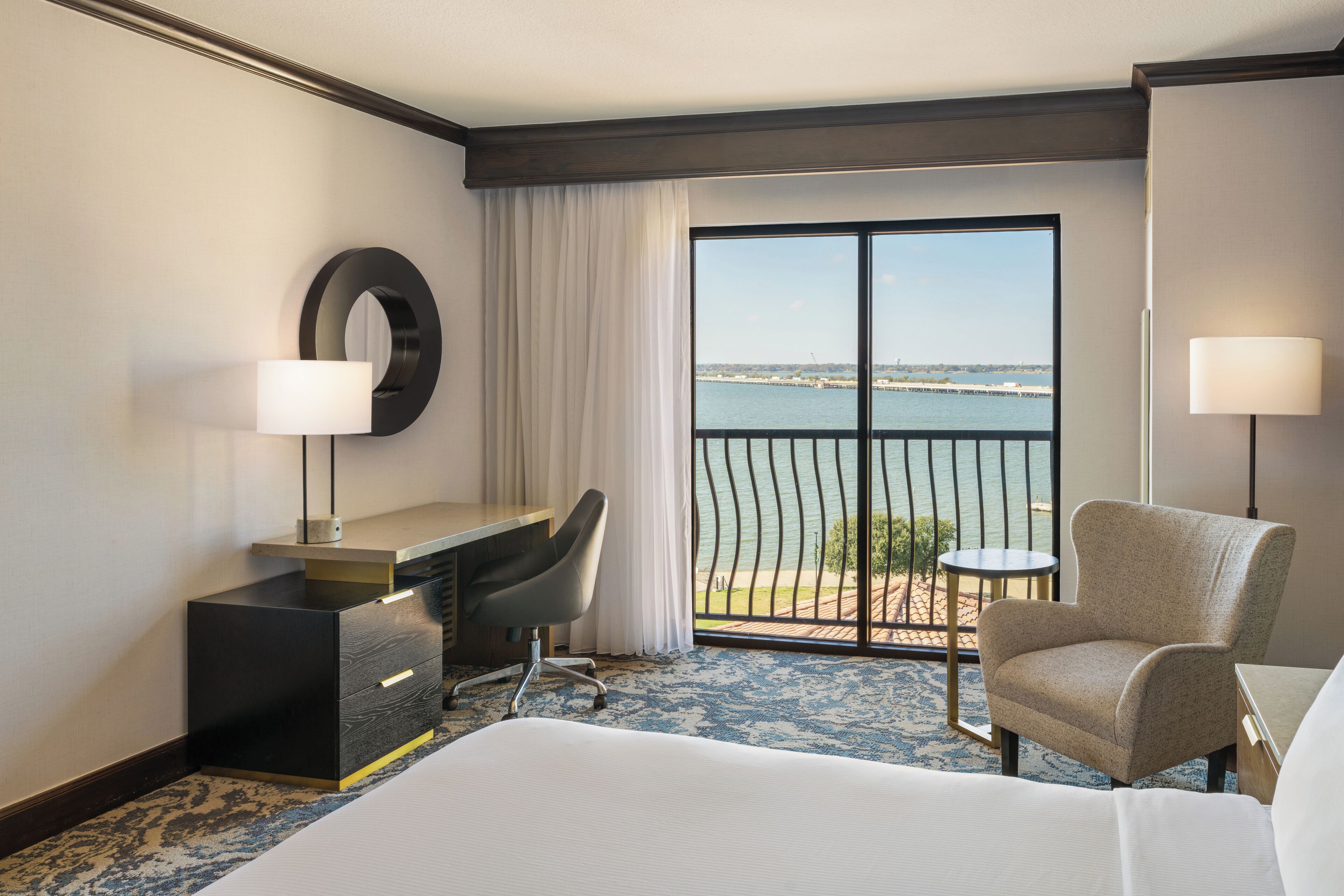 A hotel room with a bed, a desk with a chair, a small table, a lamp, and a view of the sea through a large window.