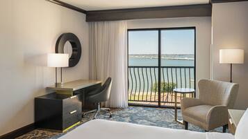 A hotel room with a bed, a desk with a chair, a small table, a lamp, and a view of the sea through a large window.