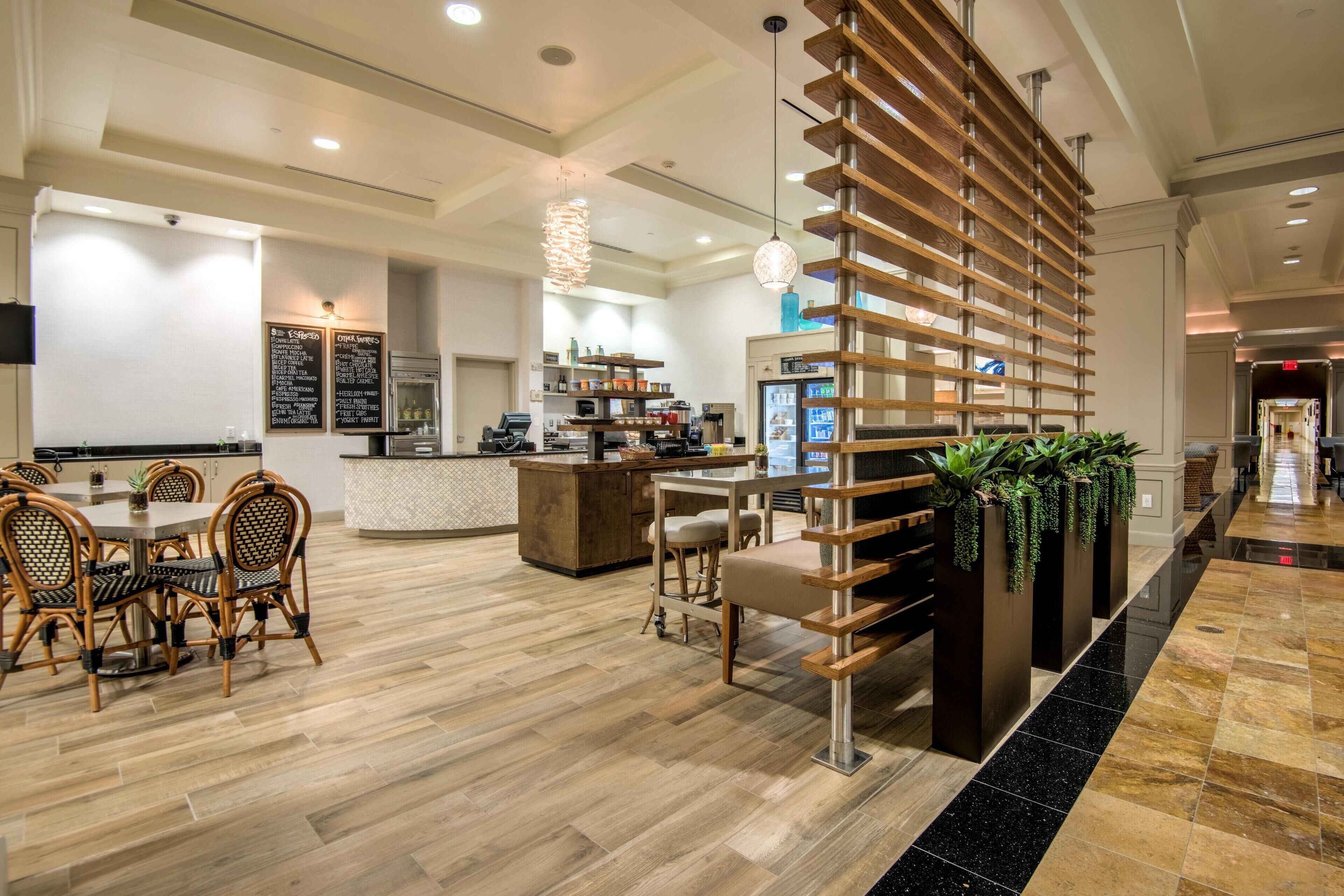 Un vestíbulo de hotel moderno con un mostrador de recepción de madera, un comedor con sillas de mimbre y una zona de bar.