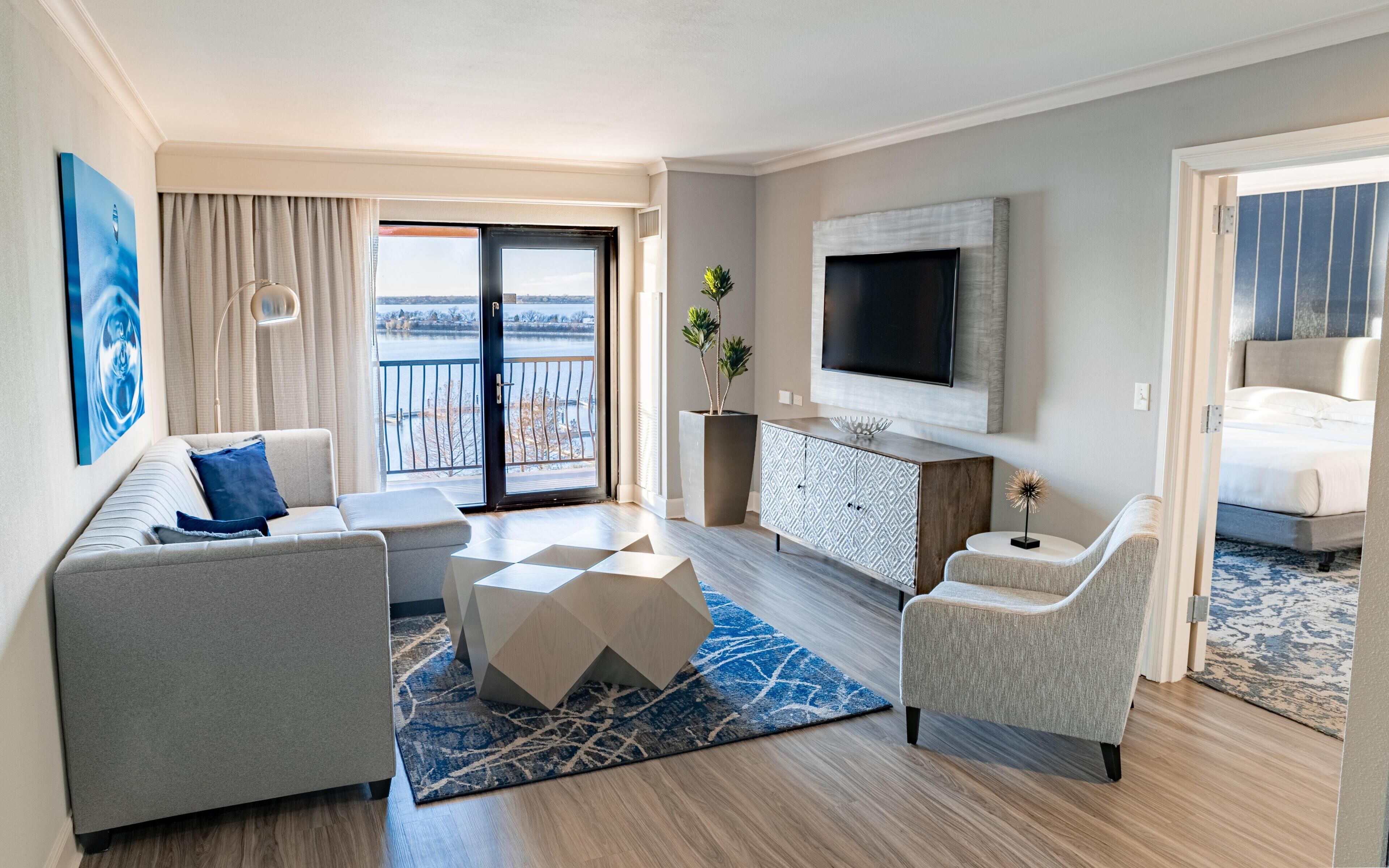 A modern hotel room with a sofa, armchair, and a flat-screen TV mounted on the wall.