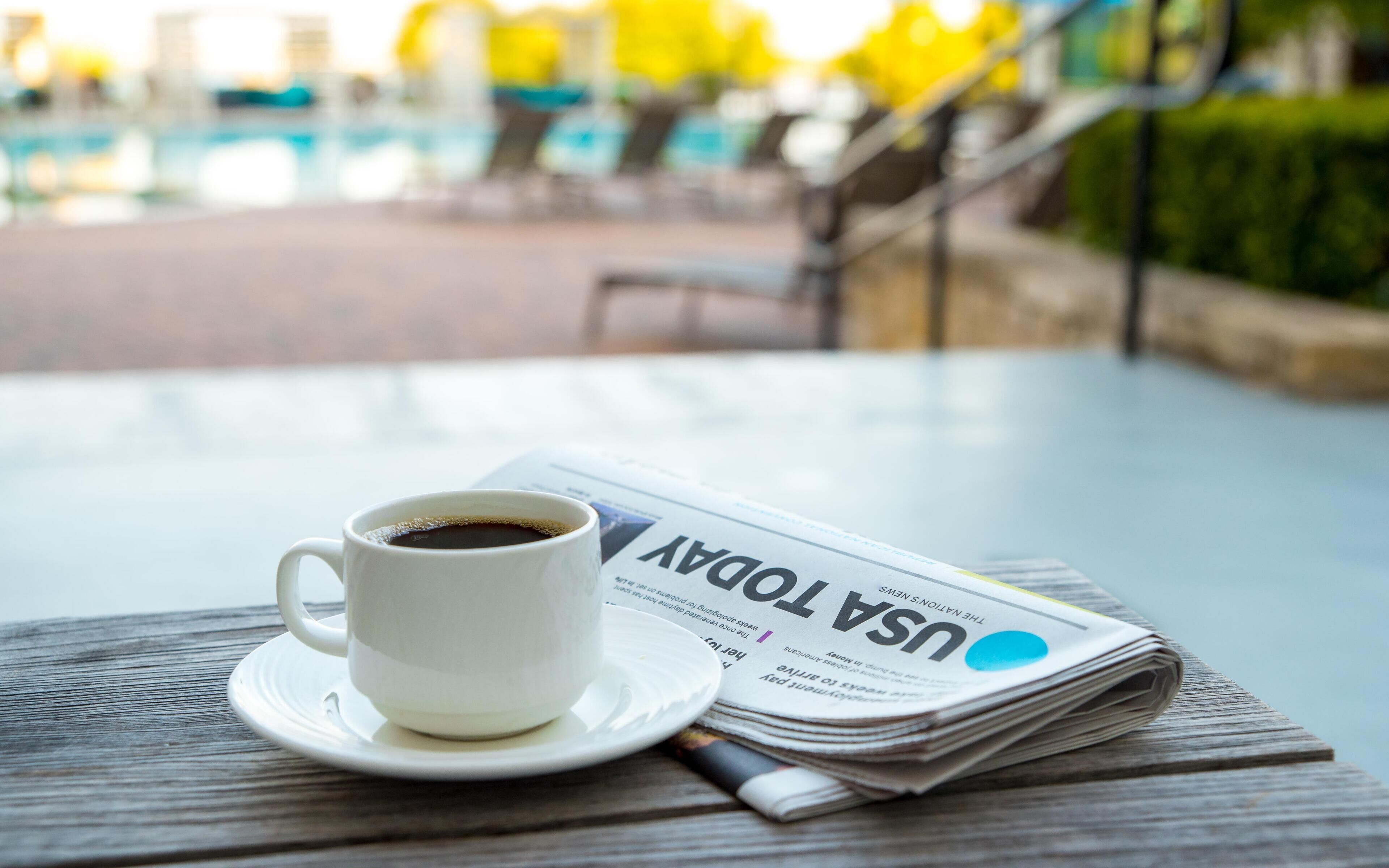 En hvid kop kaffe på en underkop, en avis med titlen 'USA TODAY', og et træbord.