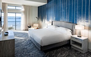 A hotel room with a large bed, two bedside tables, a desk, a chair, a view of the outdoors, and a blue-striped wall. - Hilton Dallas/Rockwall Lakefront (Rockwall)