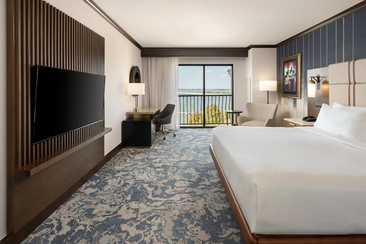 A hotel room with a large bed, a flat-screen TV, a desk with a chair, a small table, and a balcony with a view.