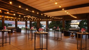 An indoor venue with multiple tables and chairs, string lights, and large windows offering a view of the outdoors.