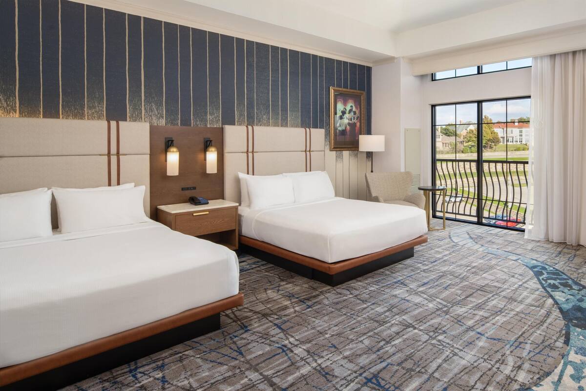 A hotel room with two beds, a large window with a view, a patterned carpet, and a painting on the wall.