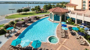 A swimming pool with lounge chairs and umbrellas, a view of the water, and a building with a red roof. - Hilton Dallas/Rockwall Lakefront (Rockwall)