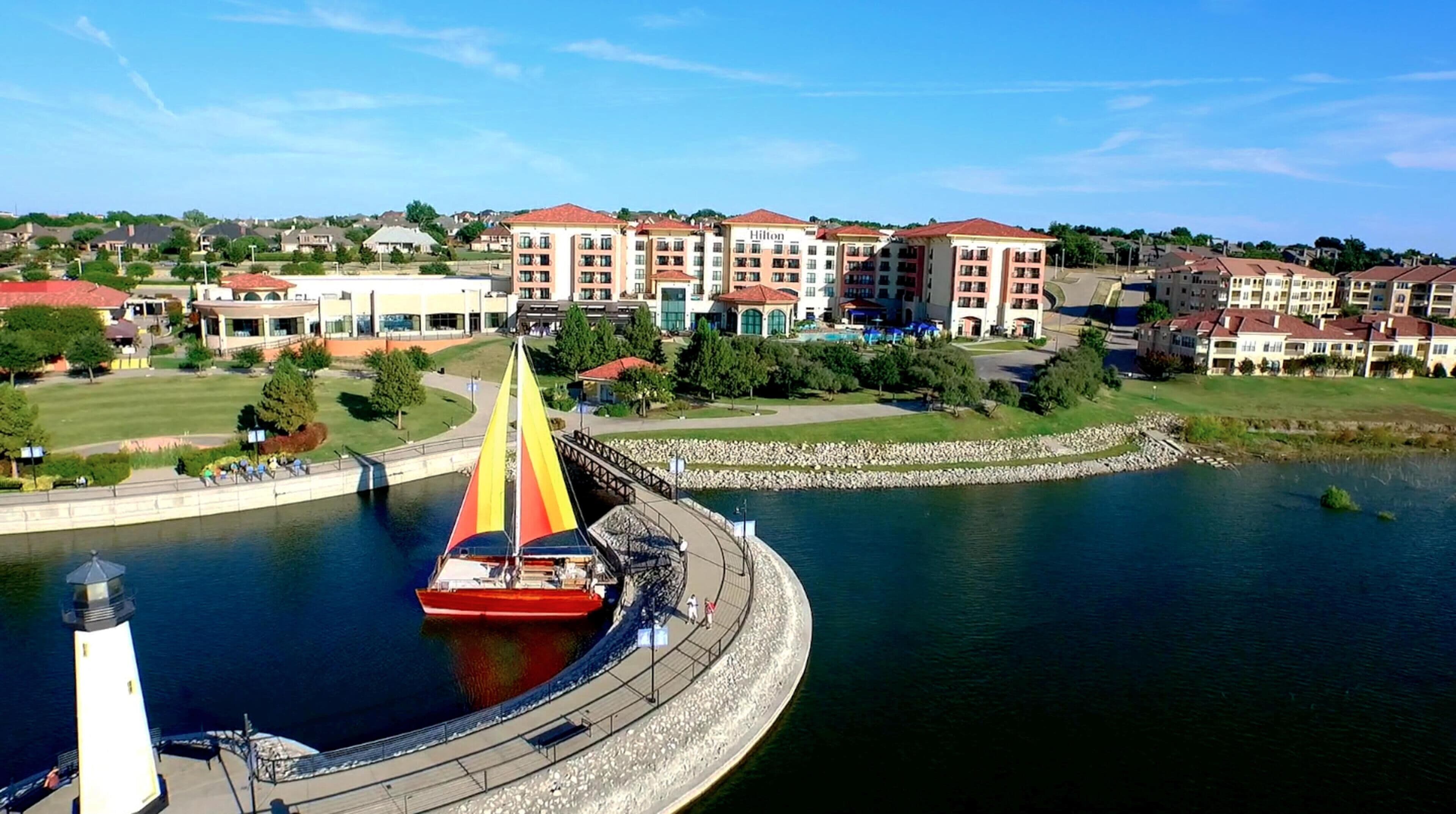 Un voilier sur un lac, avec une promenade en bord de mer, entouré d’immeubles à plusieurs étages portant l’enseigne « Hilton ».