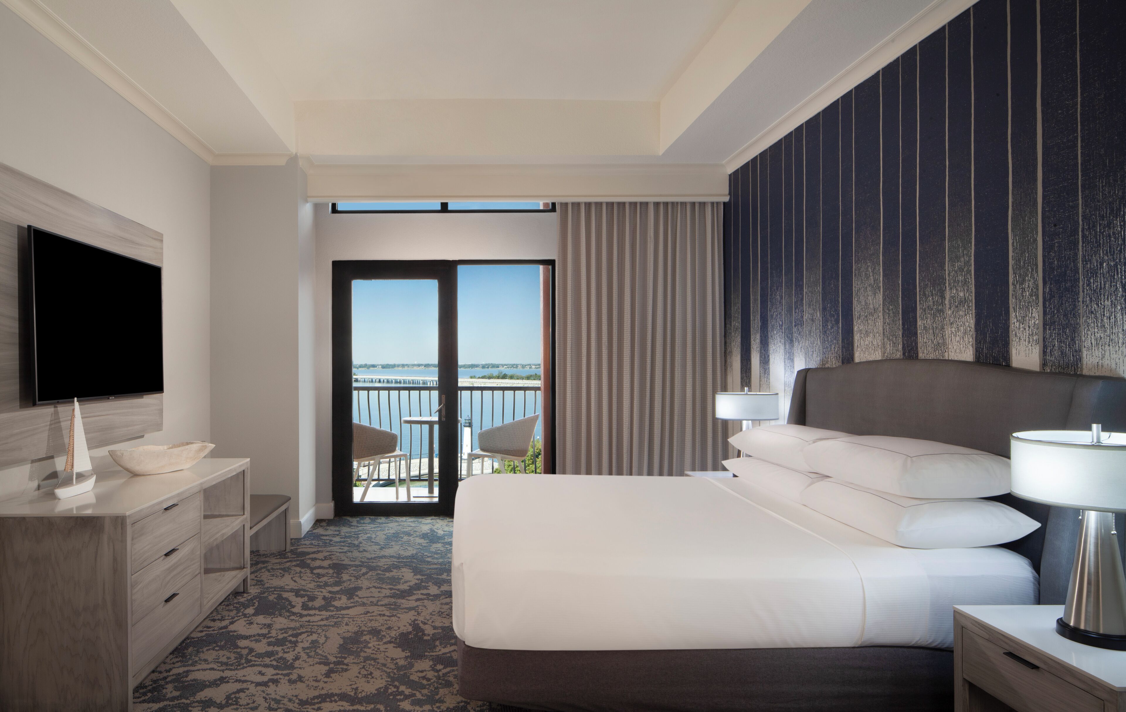 Ein Hotelzimmer mit einem großen Bett, einem Fernseher, einem Schreibtisch und Meerblick vom Balkon.