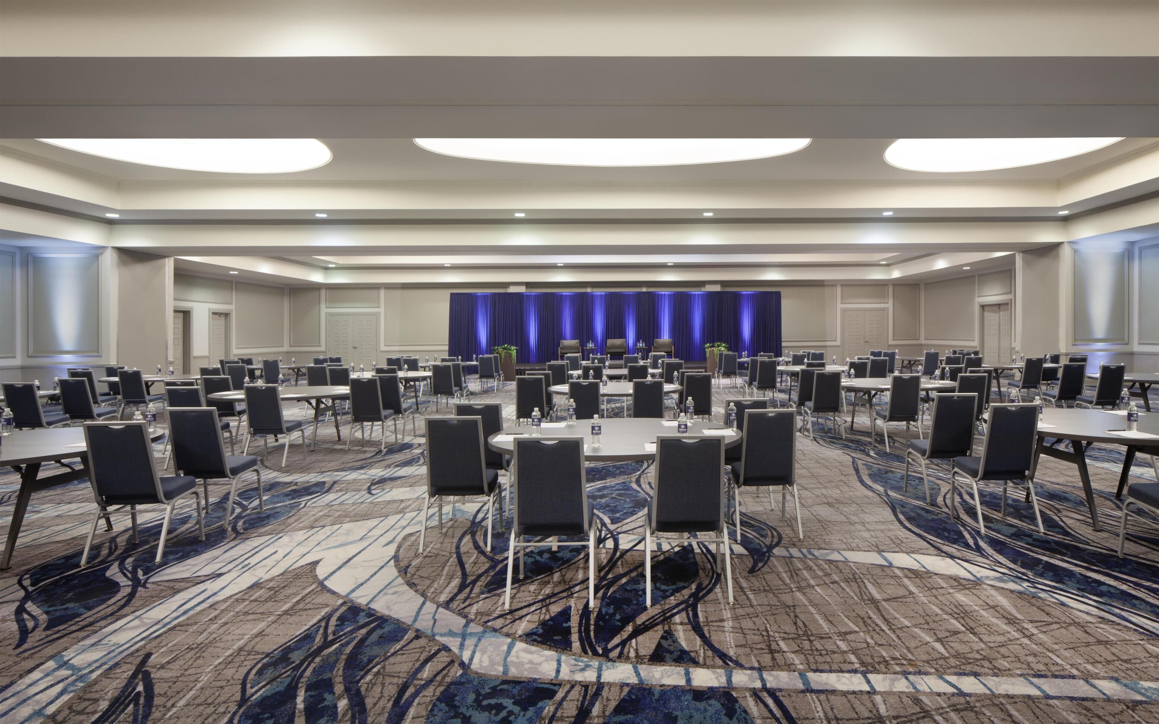 A conference room with round tables, chairs, and a stage with a blue curtain backdrop.