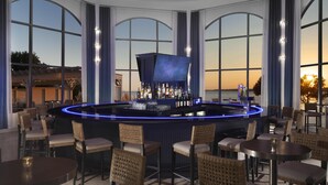 A bar area with a curved counter, bar stools, and a view of the sunset through large windows.