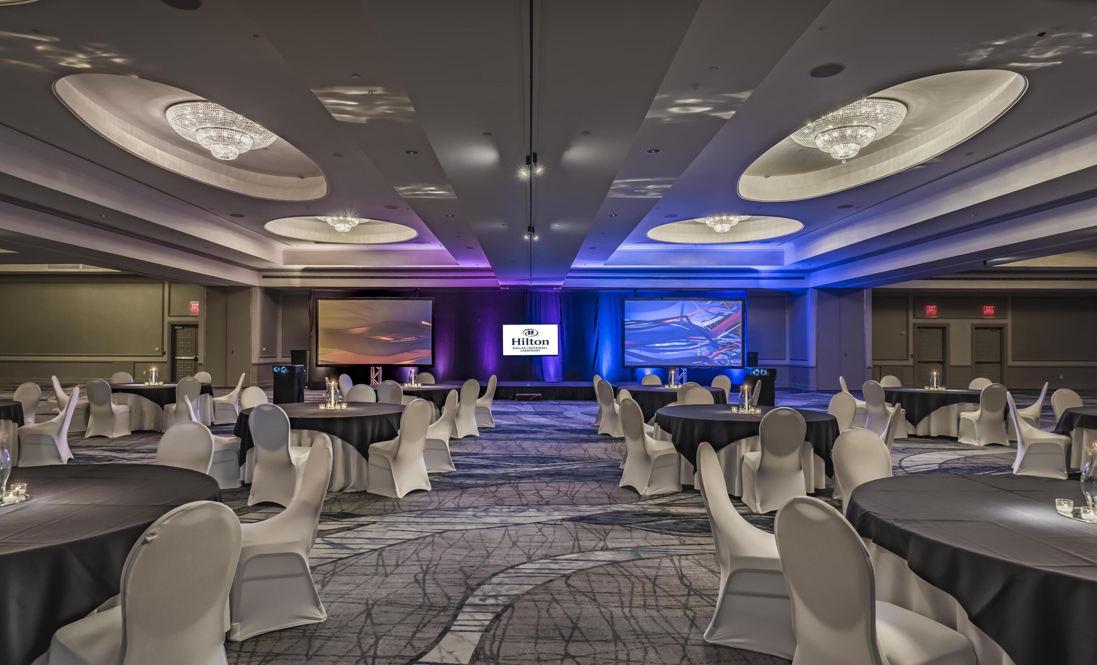 Une salle de conférence avec des tables rondes, des chaises blanches, un grand écran et le logo de Hilton.