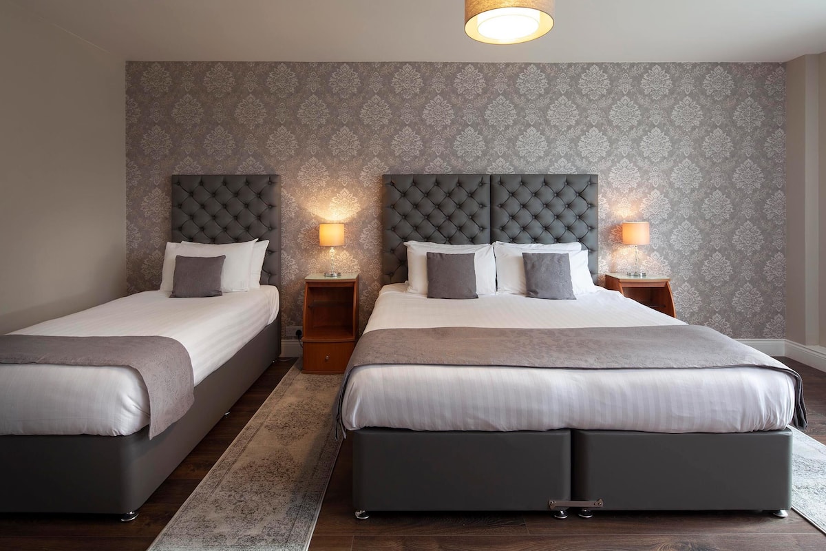 A hotel room with two beds, each with a grey headboard and white bedding. There are bedside tables with lamps, and a patterned wallpaper on the walls.