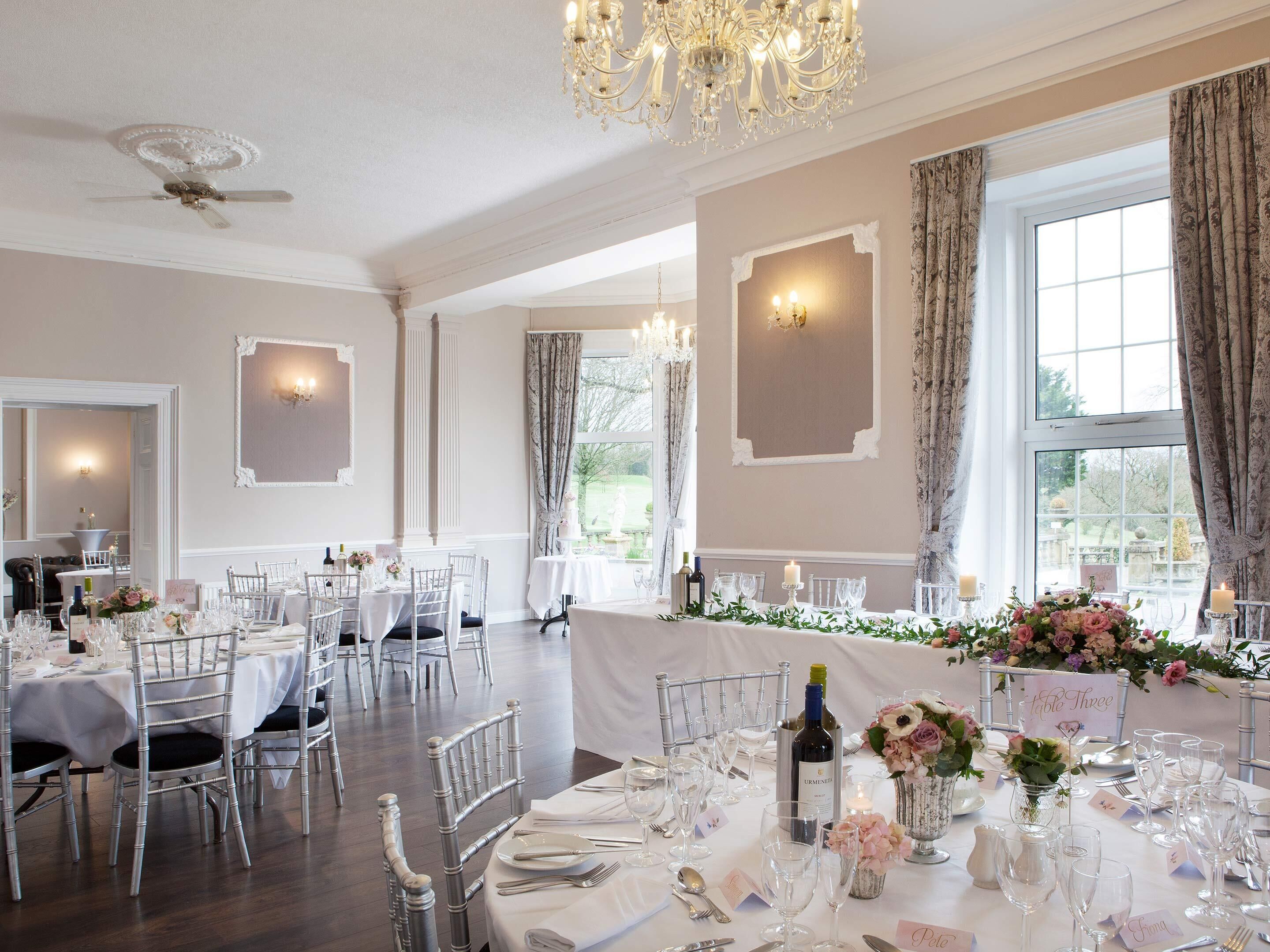 Comedor formal con varias mesas dispuestas para una comida, candelabros elegantes y amplias ventanas con cortinas.