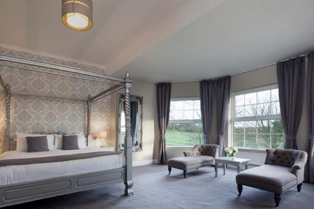 A four-poster bed with a canopy, a chaise lounge, a small table, and a view of the outdoors through large windows.