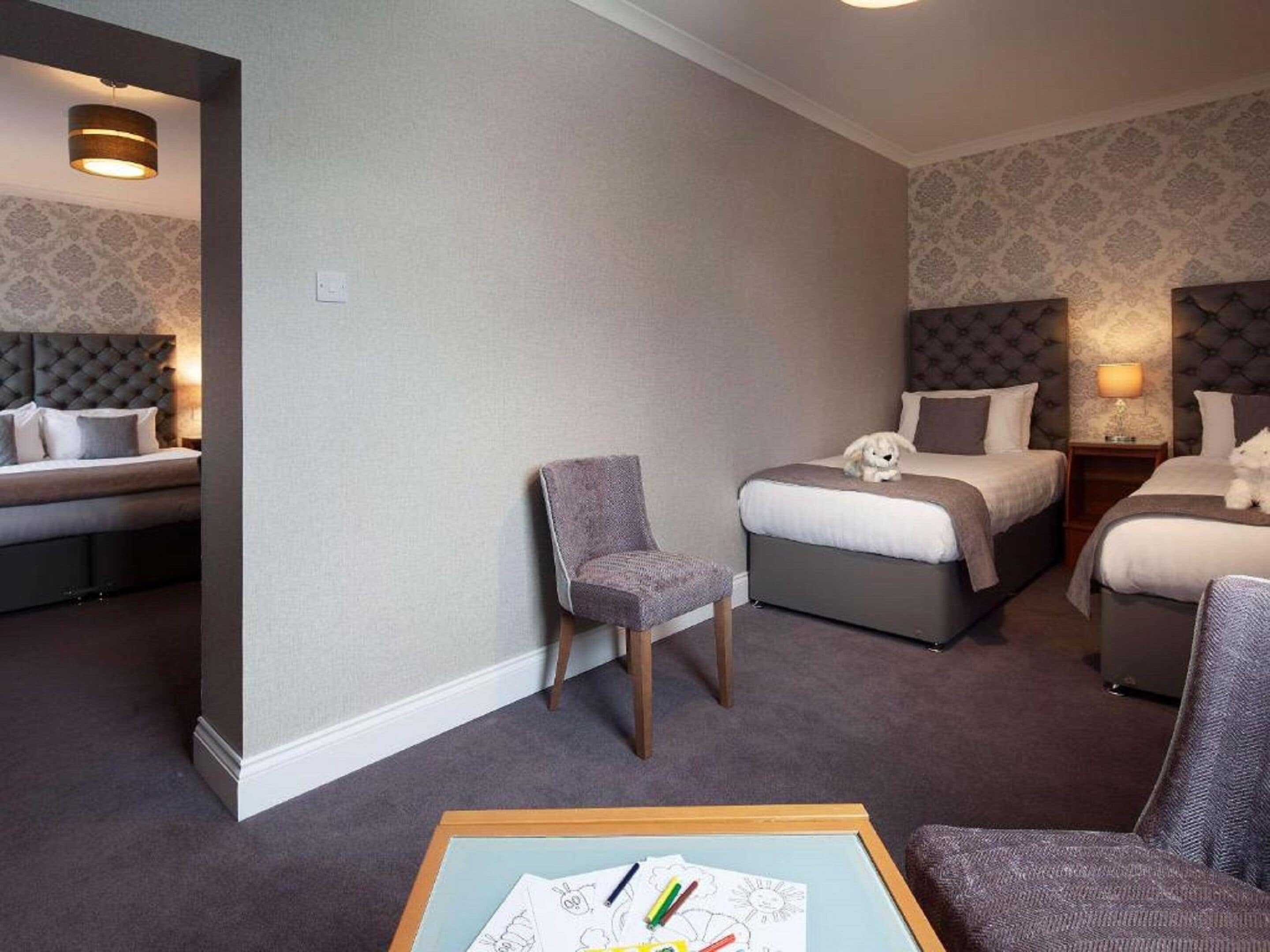 A hotel room with a bed, a chair, a small table with papers, and a nightstand with a lamp.