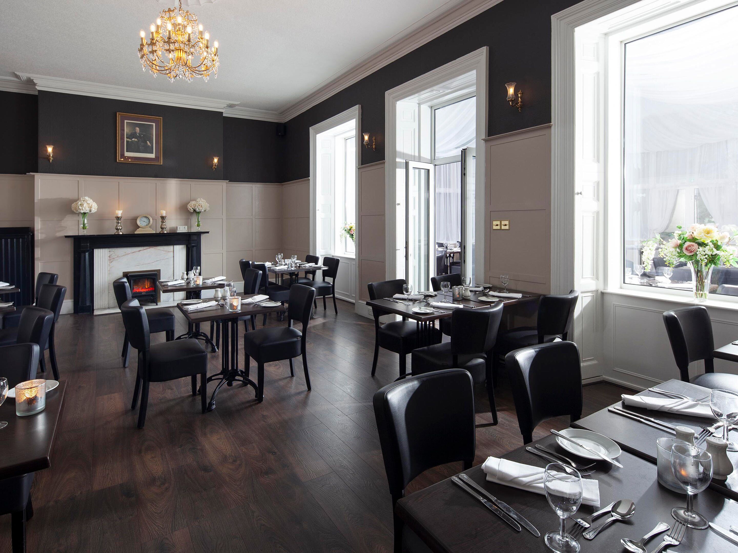 A dining area with dark wood flooring, black chairs, and tables set for a meal. There is a fireplace with a fire, and a chandelier hanging from the ceiling.