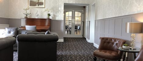 Un hall de hotel con una alfombra estampada, sofás de cuero, un sillón de cuero marrón y una mesita con una lámpara.