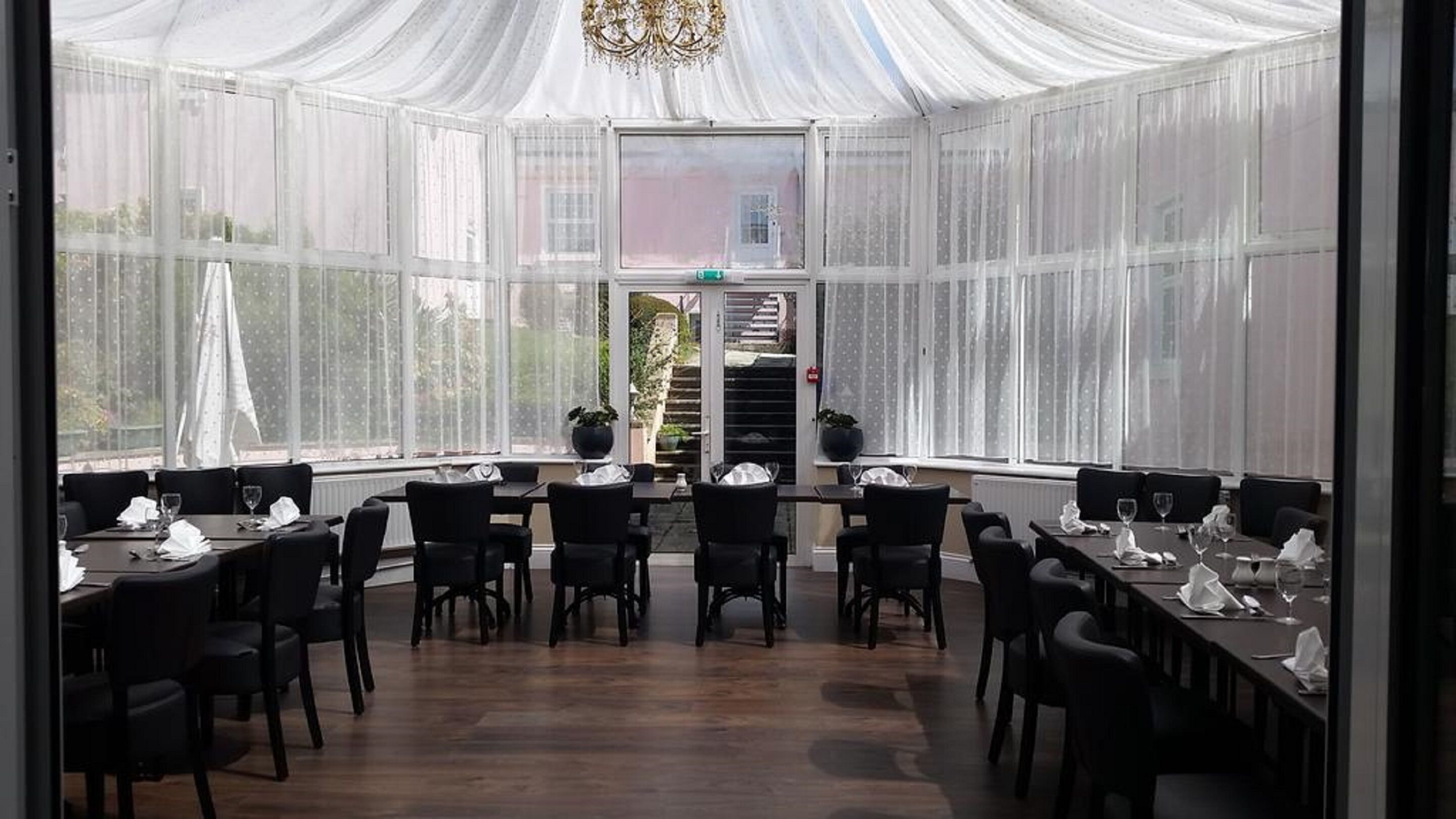 Une grande pièce, bien éclairée, avec un plafond haut et de longues draperies blanches et transparentes. Des tables sont disposées, accompagnées de chaises et de verres à vin, ce qui indique qu’il s’agit d’un espace dédié aux repas.