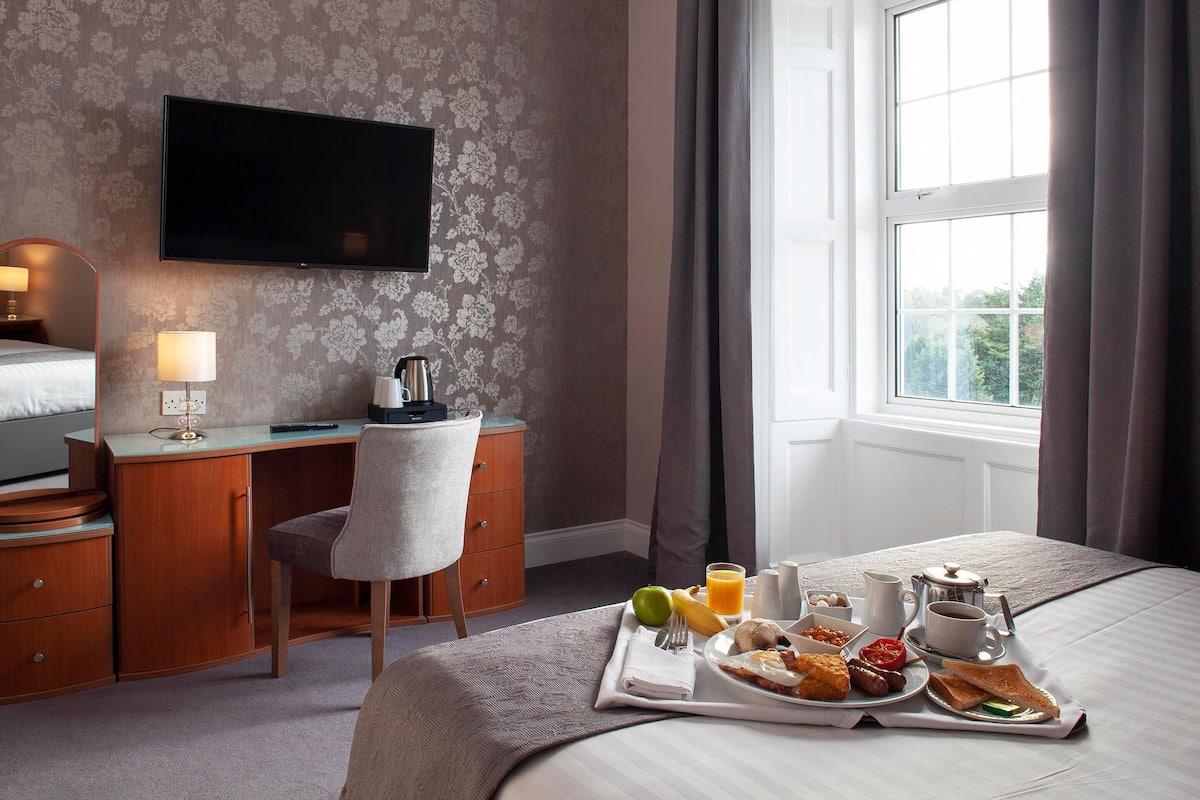 A hotel room with a bed, a desk with a chair, a TV, and a breakfast tray with eggs, sausages, toast, and juice.