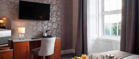 A hotel room with a bed, a desk with a chair, a TV, and a breakfast tray with eggs, sausages, toast, and juice.