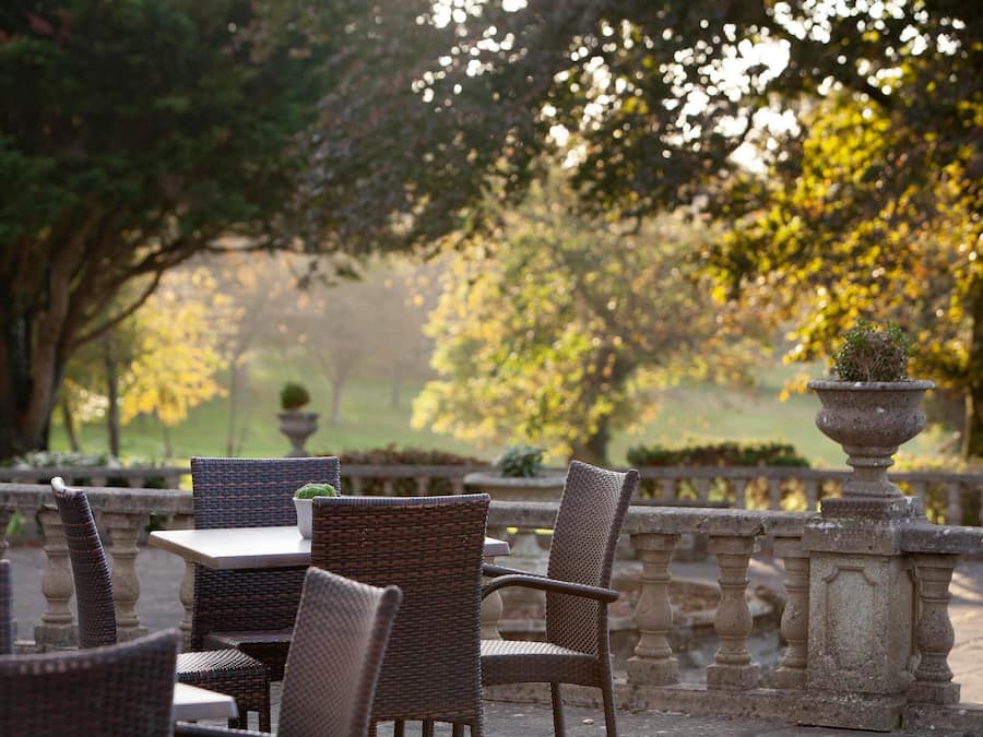 An outdoor seating area with wicker chairs and tables, surrounded by stone balustrades and greenery.