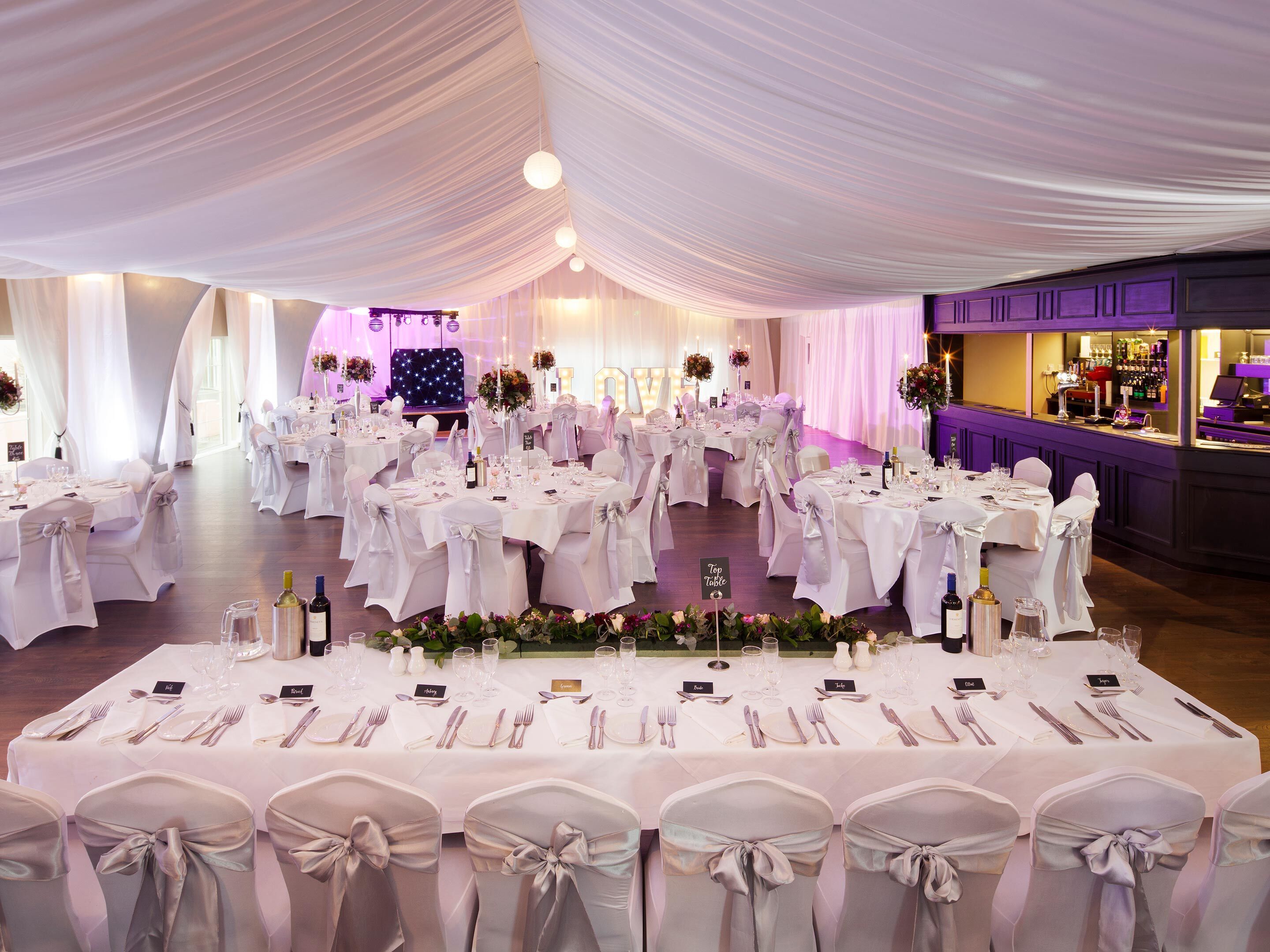 A banquet hall with round tables set for a formal event, featuring white tablecloths, silverware, and floral centerpieces.