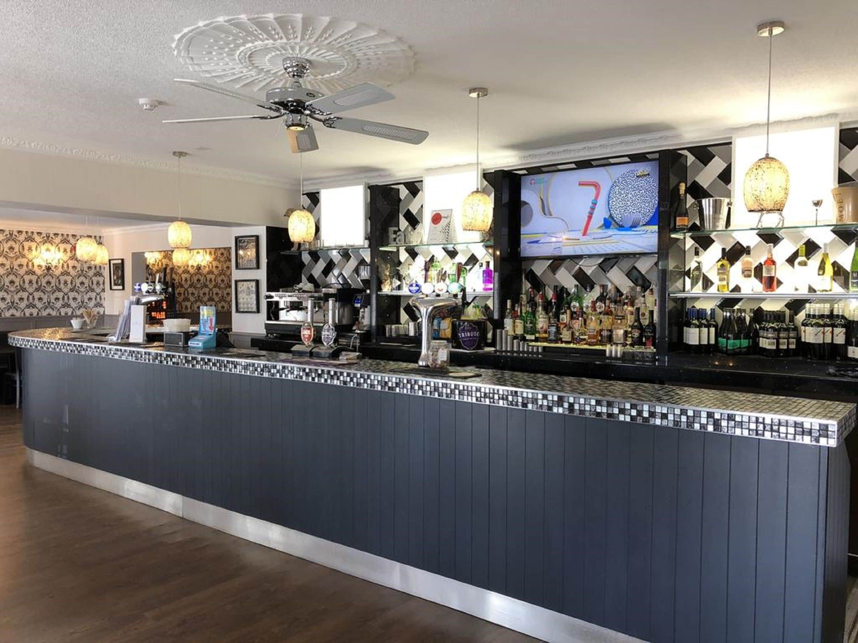 A bar area with a counter, bar stools, a ceiling fan, and a television screen displaying a sports game.
