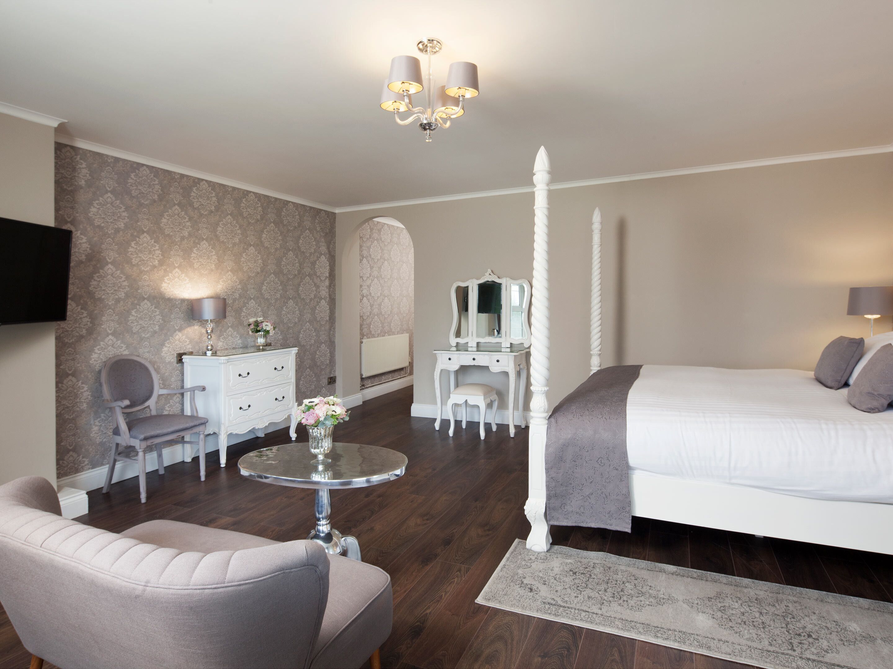 Een slaapkamer met een hemelbed, een kaptafel met spiegel, een stoel, een klein rond tafeltje met een bloevas en een aan de muur bevestigde televisie.