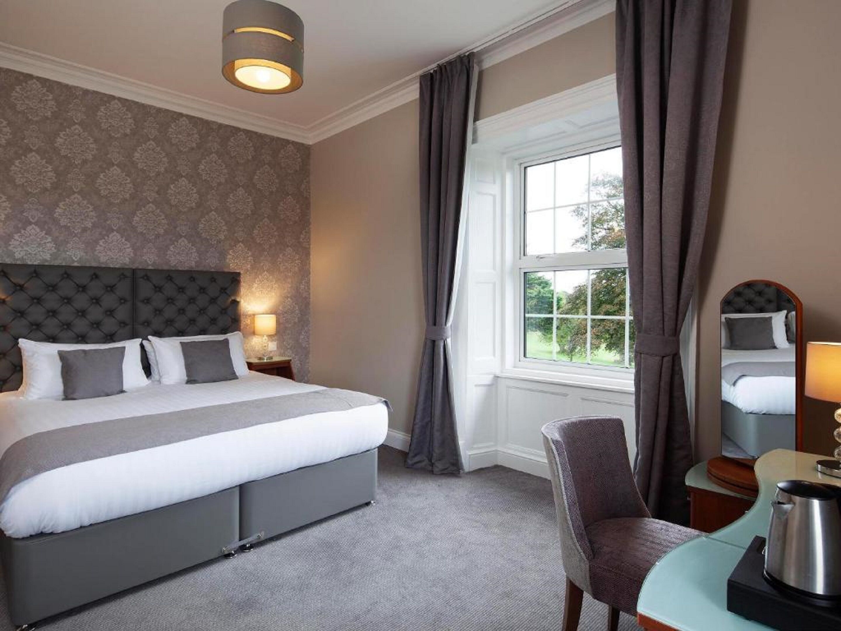 A hotel room with a large bed, a chair, a desk with a kettle, and a window with curtains.