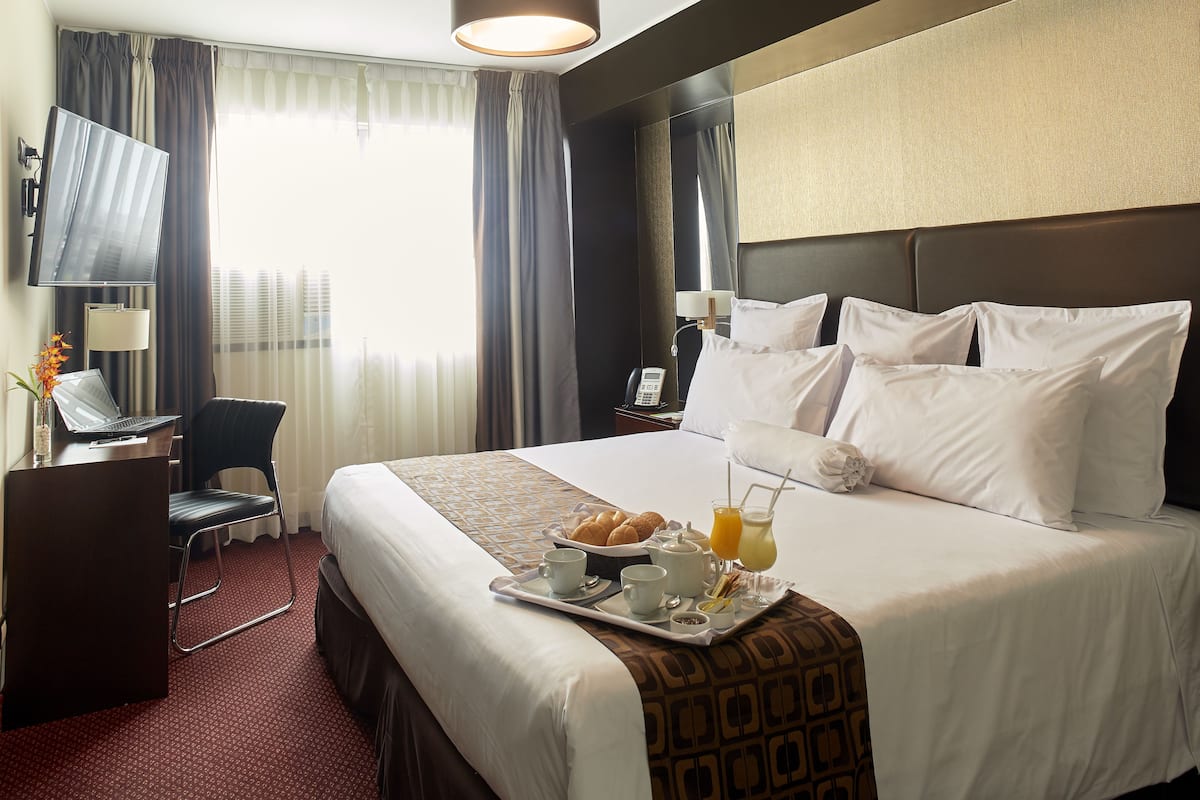 Quarto de hotel com uma cama, uma escrivaninha com um laptop, uma cadeira, uma bandeja com itens de café da manhã e uma mesa de cabeceira com luminária e telefone.